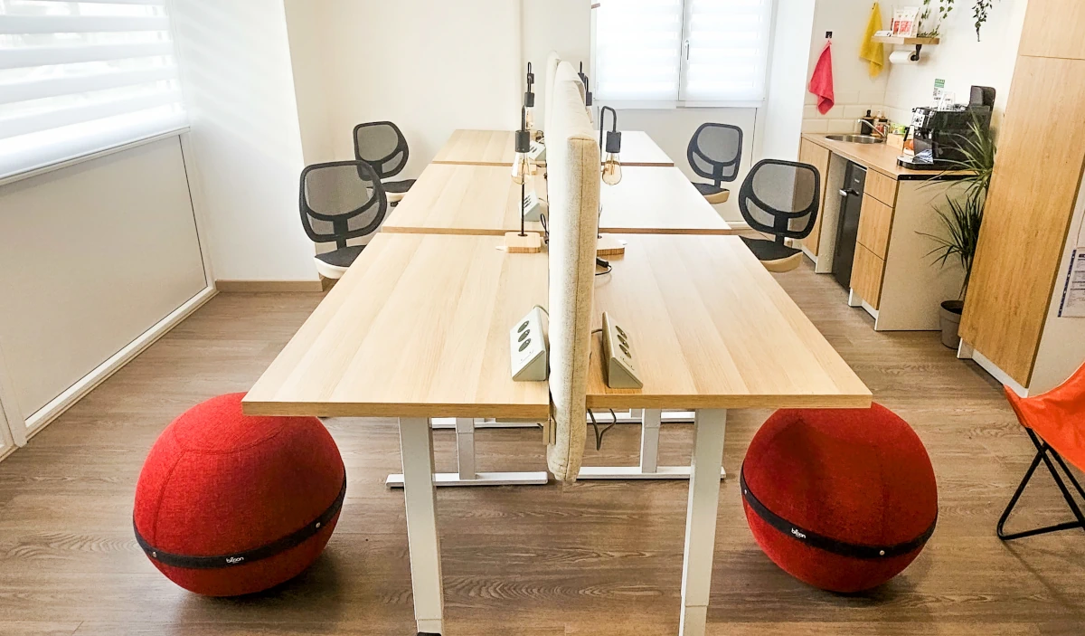 Photo de l'espace de coworking mettant en avant les deux rangées de trois postes:
                        l'espace est dégagé et lumineux, le plancher en bois, les murs blancs et les bureaux
                         sont ajustables en hauteurs. Deux sont équipés de ballon d'assise ergonomiques, et
                         quatre sont équipé d'office bike, des chaises de bureau où on pédale.